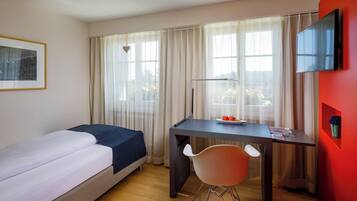 Une chambre d’hôtel comprenant un lit, un bureau avec une chaise, un téléviseur fixé au mur et une fenêtre avec des rideaux.