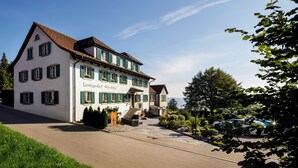 A two-story building with a white facade and green shutters, labeled "Landgasthof Woselberg." It features a front entrance with steps, a driveway, and a parking area with cars.