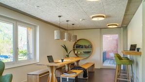 Un'area di lavoro moderna con un tavolo in legno, panche e un computer portatile. Sono presenti scaffali e una grande finestra che si affaccia su vegetazione e un edificio.