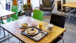 Ein Frühstückswagen mit Toast, Obst und Getränken auf einem Holztisch in einem modernen Essbereich mit grünen Stühlen und einem grauen Sessel.