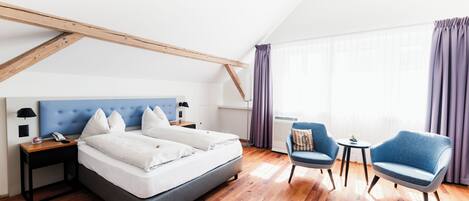 Ein modernes Hotelzimmer mit einem großen Bett, zwei Sesseln, einem kleinen Tisch und einem Fenster mit Vorhängen.