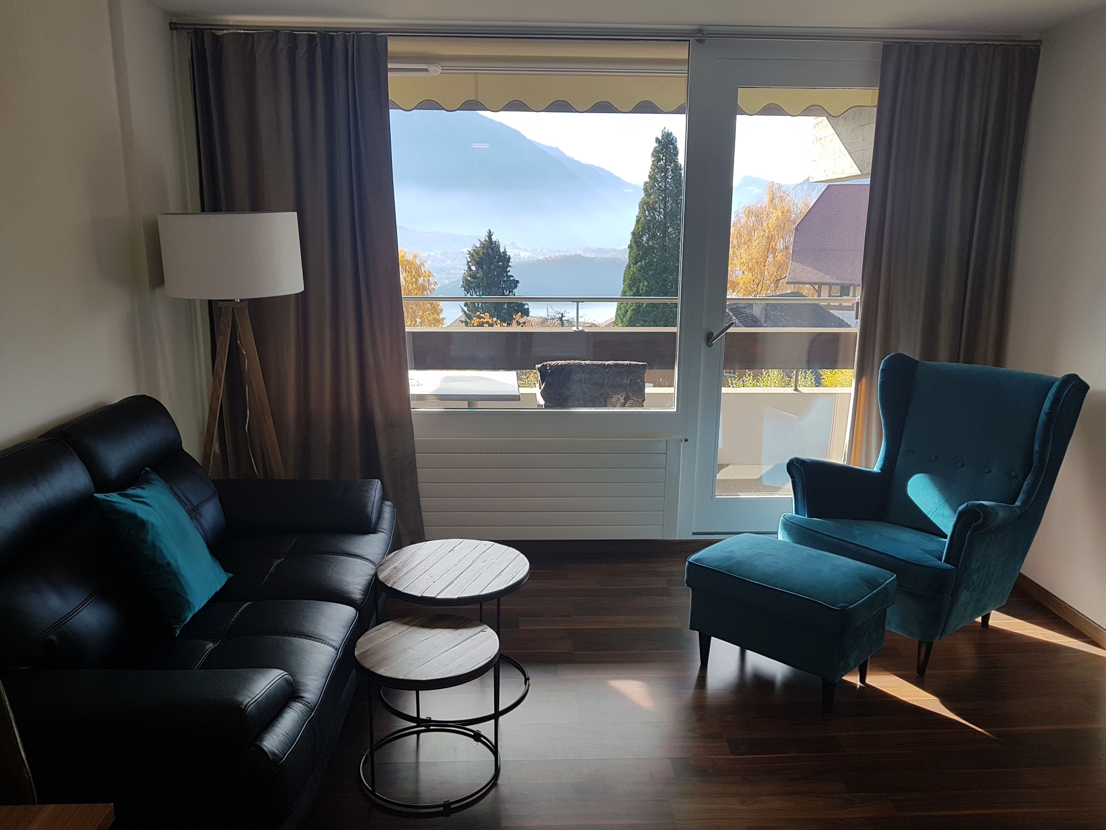 A modern living room with a black leather sofa, a blue armchair, and a small wooden table. A large window offers a view of mountains and a balcony.