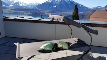 Ein Balkon mit Stuhl und Kissen mit Blick auf eine Berglandschaft mit See und Kirche.