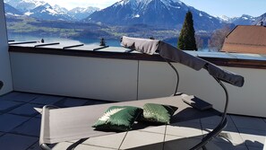 A balcony with a chair and a cushion, overlooking a mountainous landscape with a lake and a church.