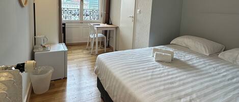 A hotel room with a bed, a desk, a chair, a small table, a trash bin, and a window with a view of buildings.