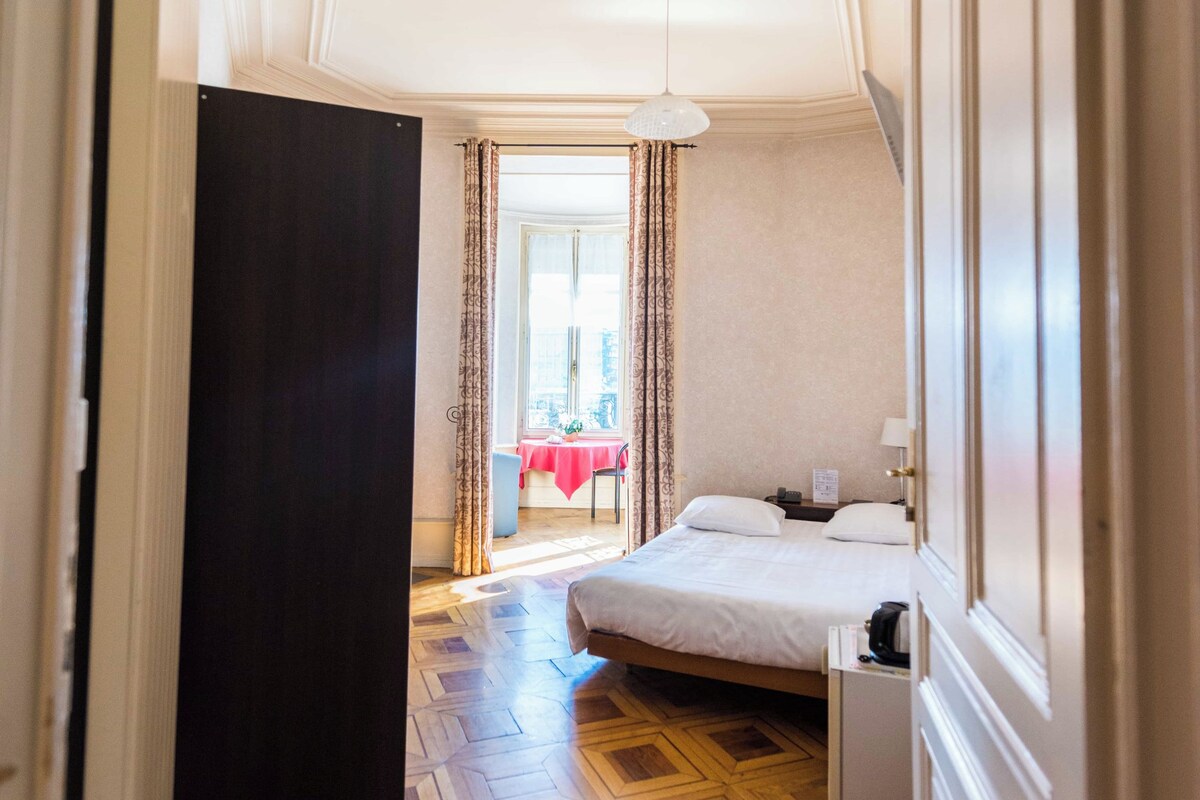 Habitación de hotel con cama, mesita de noche, puerta y vista a un comedor.