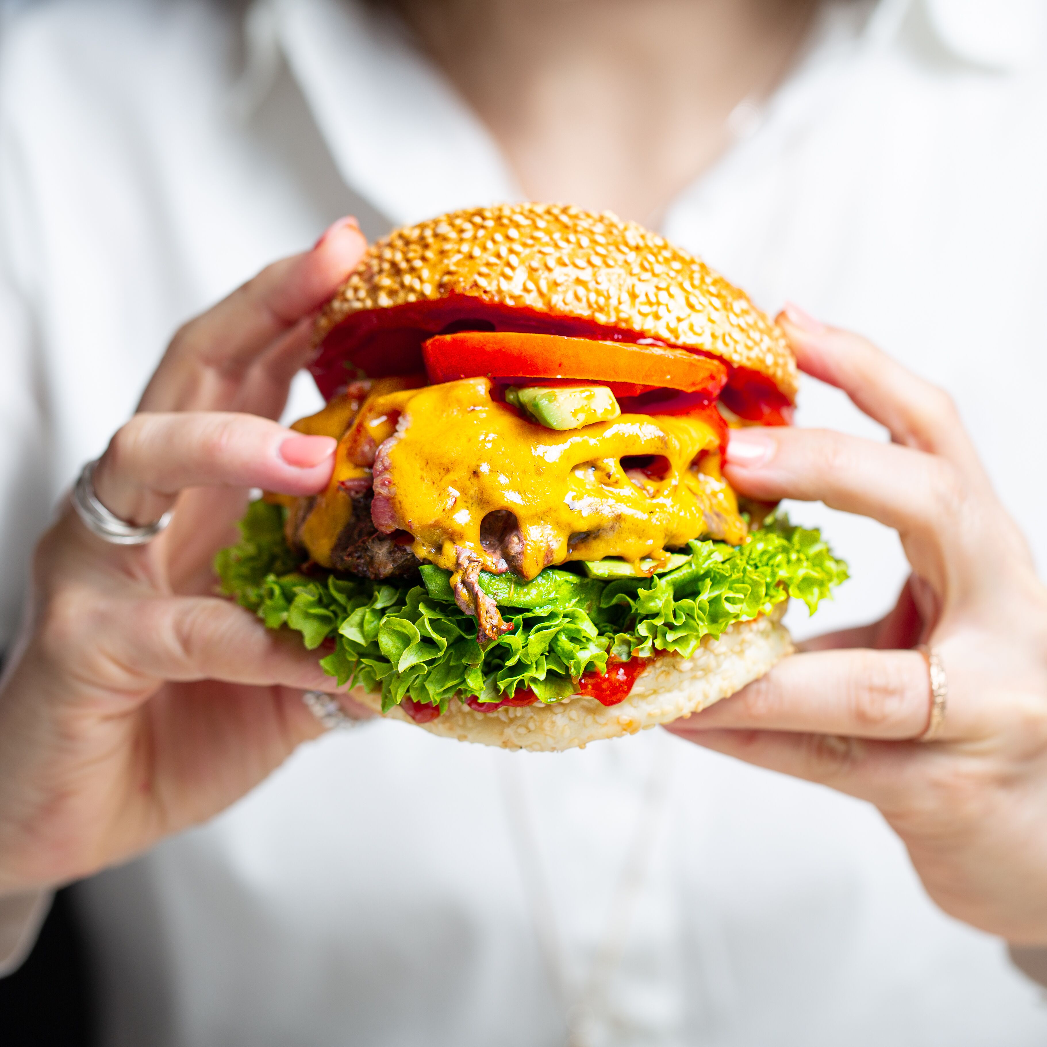 A person holding a large hamburger with visible ingredients like lettuce, tomato, cheese, and a sesame seed bun.