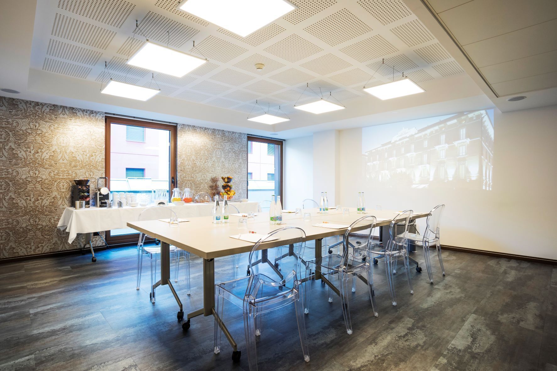 A conference room with a long table, clear chairs, a projector screen, and a patterned wall.