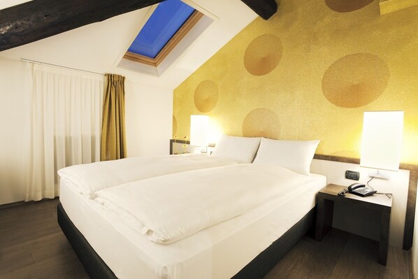 Quarto de hotel com uma cama grande, duas mesas de cabeceira com abajures, um claraboia e uma parede estampada.