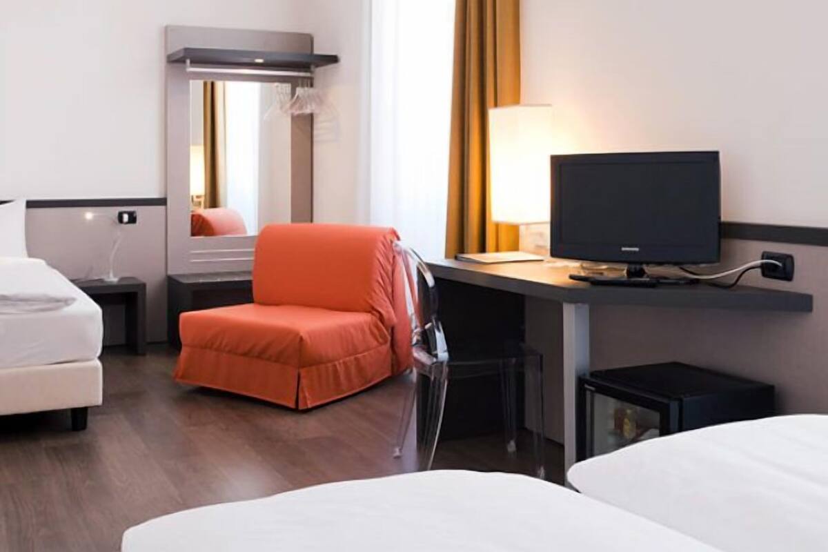 A hotel room with a bed, a desk with a computer, a chair, a mirror, and a window with curtains.
