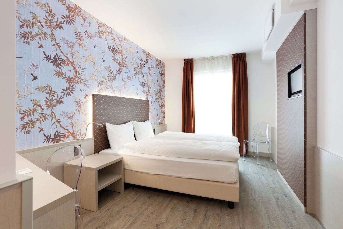 A modern hotel room with a large bed, a bedside table, a mirror, and a window with curtains.