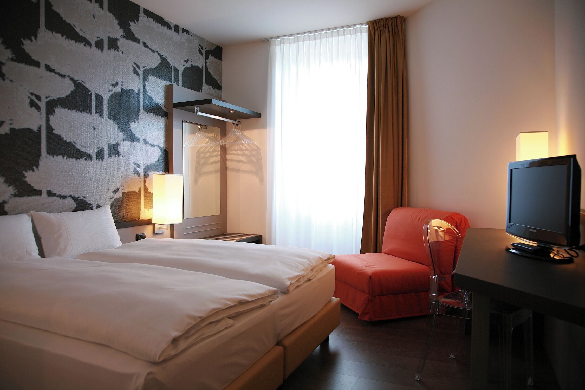 Ein Hotelzimmer mit einem großen Bett, einem Schreibtisch mit Fernseher, einem Sessel und einem Fenster mit Vorhängen.