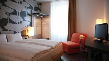 Ein Hotelzimmer mit einem großen Bett, einem Schreibtisch mit Fernseher, einem Sessel und einem Fenster mit Vorhängen.