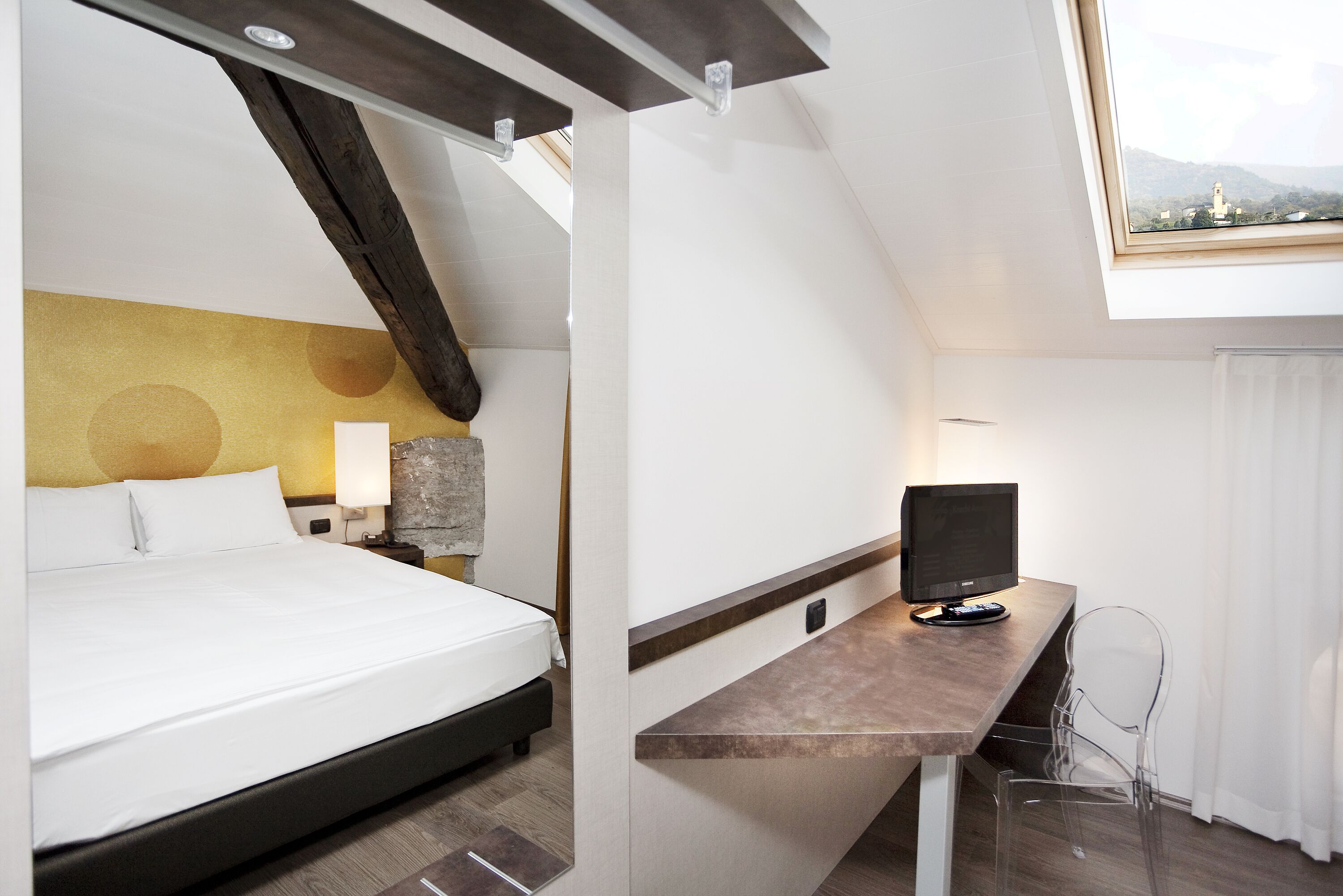Habitación de hotel moderna con cama, escritorio con computadora, lámpara y vista a las montañas a través de un tragaluz.