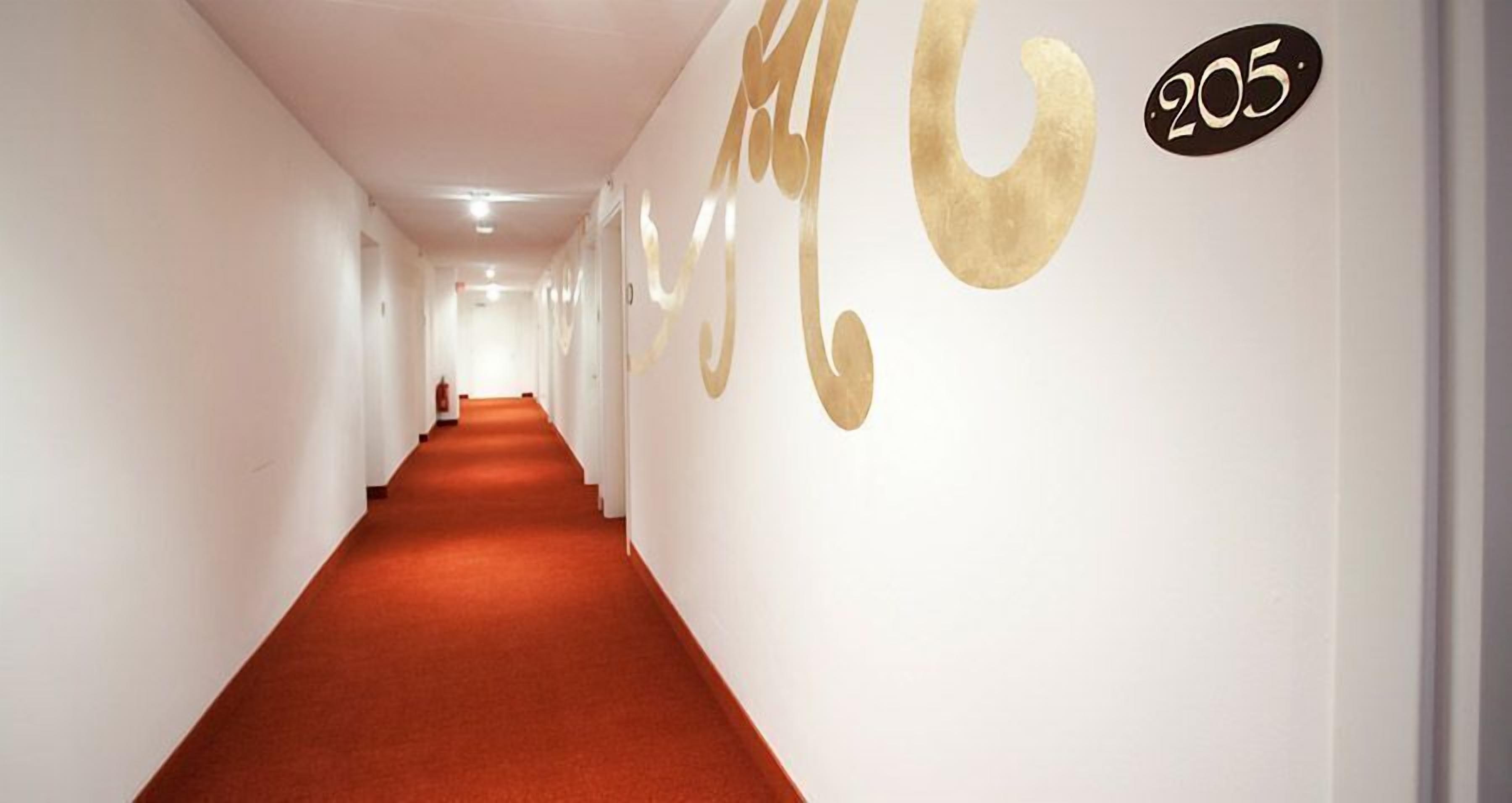 Un long couloir étroit, doté d’un tapis rouge, de murs blancs et d’un grand chiffre doré « 205 » sur le mur de droite.