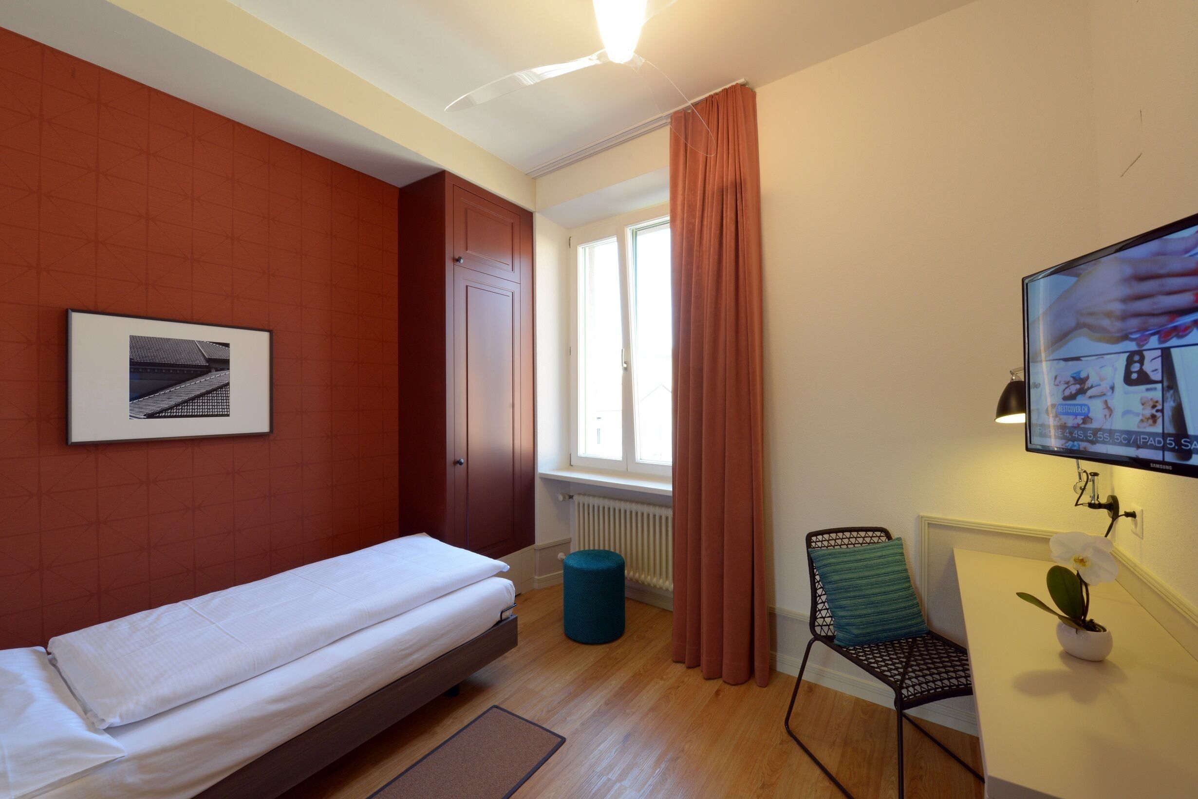Une chambre d’hôtel avec un lit, une chaise, une petite table avec une plante, une télévision et une fenêtre avec des rideaux.