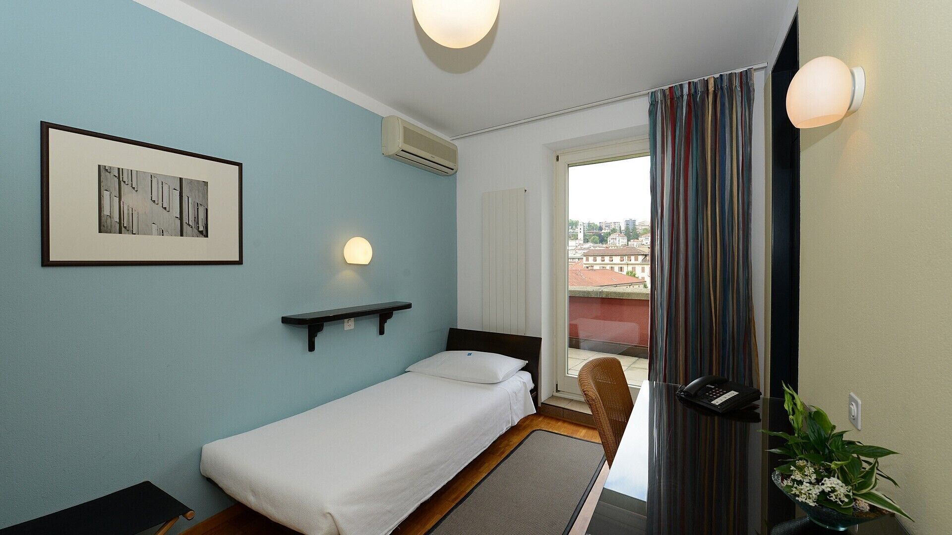 A single bed with a headboard, a bedside table, a wall-mounted light, a framed picture, an air conditioning unit, a window with curtains, and a view of buildings outside.