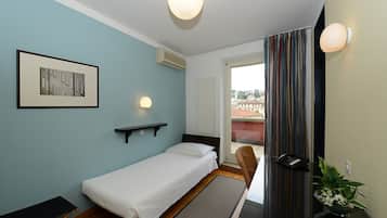 A single bed with a headboard, a bedside table, a wall-mounted light, a framed picture, an air conditioning unit, a window with curtains, and a view of buildings outside.
