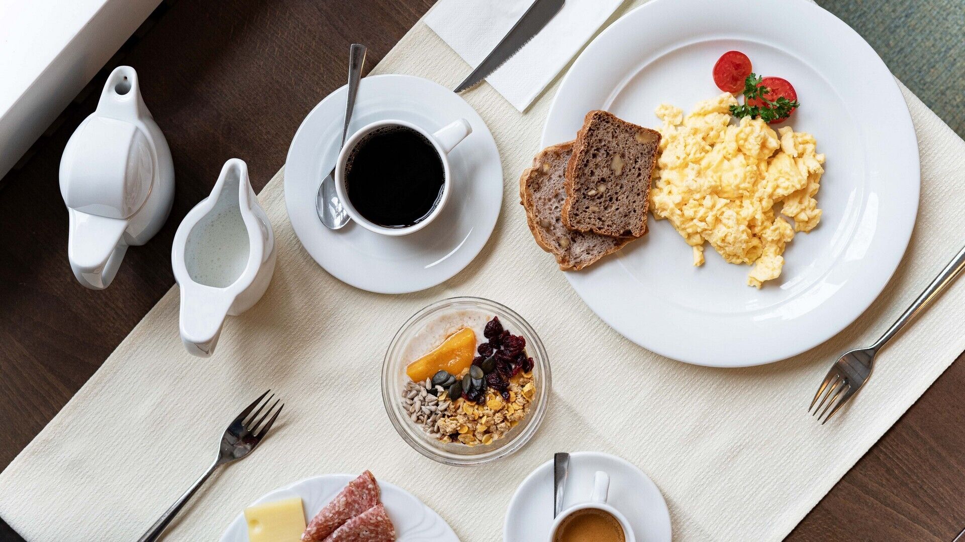 A breakfast setting with scrambled eggs, toast, a cup of coffee, a bowl of cereal, and a small plate of cheese and salami.