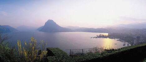 A panoramic view of a lake with a prominent mountain in the background, surrounded by greenery and a distant cityscape.