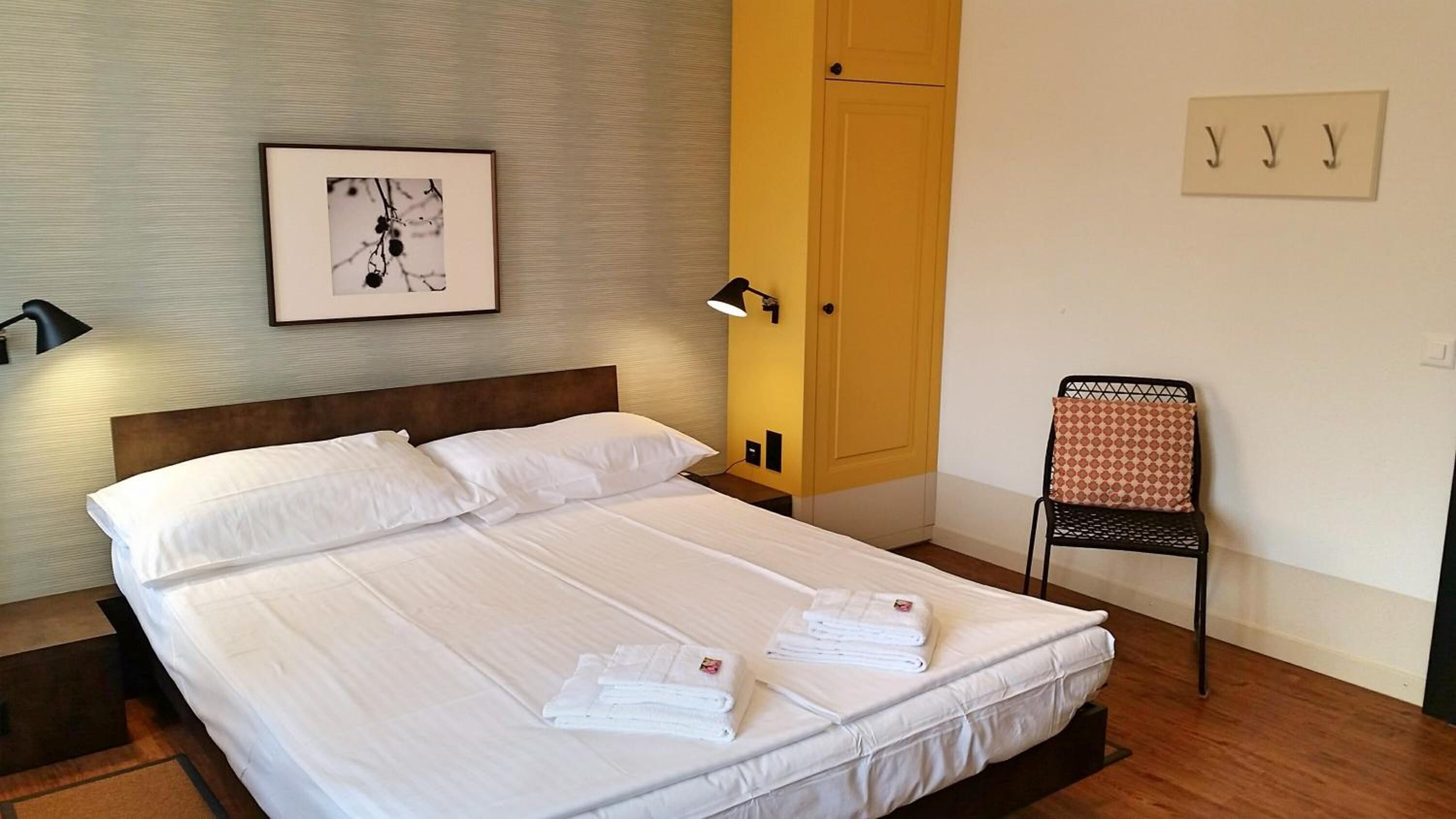 A neatly made bed with white linens and towels, a bedside lamp, a framed picture on the wall, and a chair with a patterned cushion.