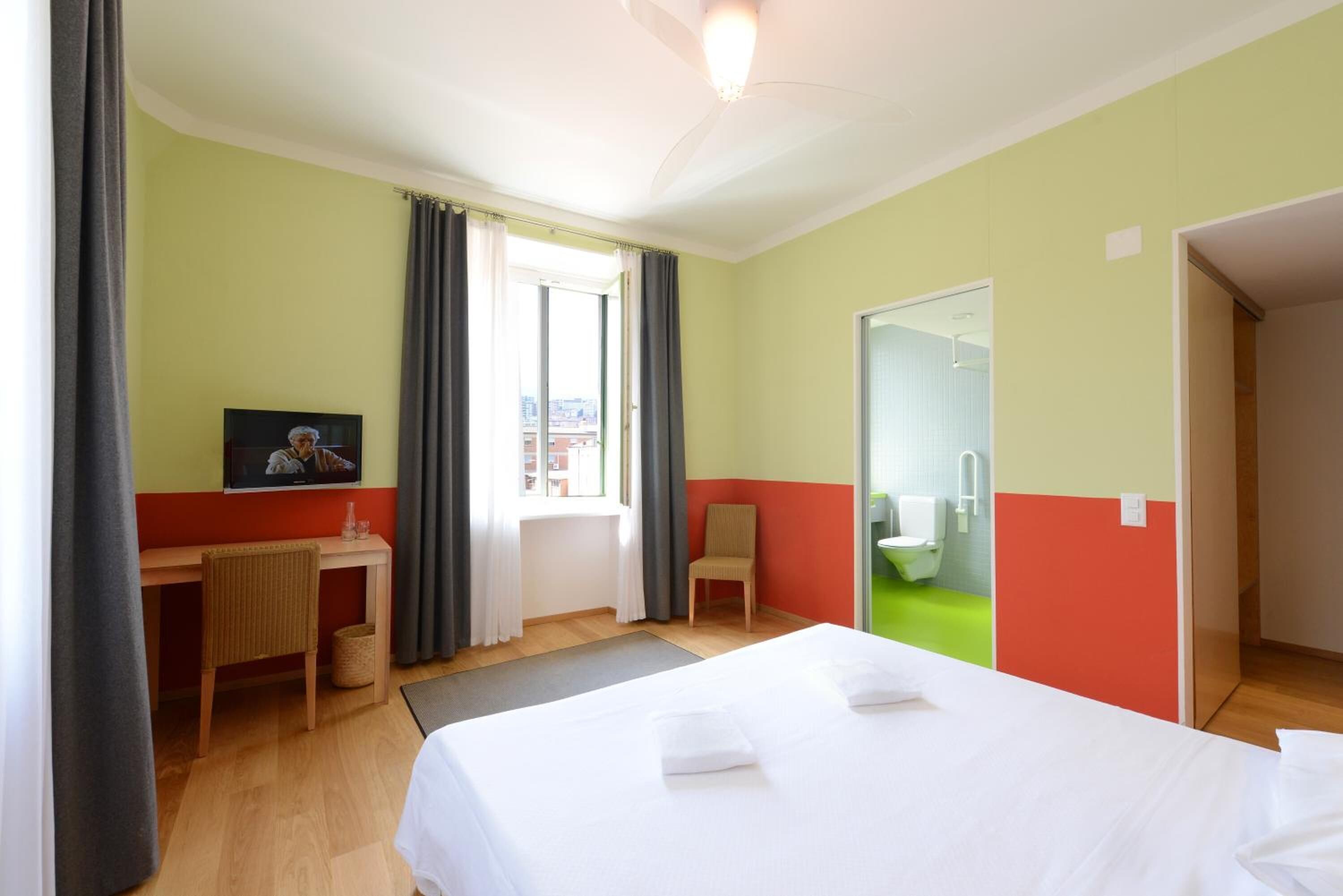 Une chambre d’hôtel avec un lit, un bureau, une chaise, une fenêtre avec des rideaux et une salle de bain visible à travers une porte ouverte.