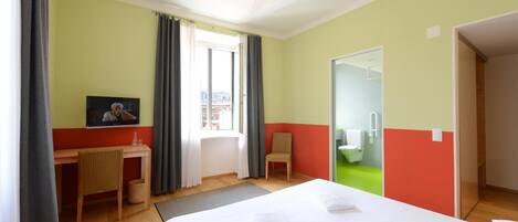 Une chambre d’hôtel avec un lit, un bureau, une chaise, une fenêtre avec des rideaux et une salle de bain visible à travers une porte ouverte.