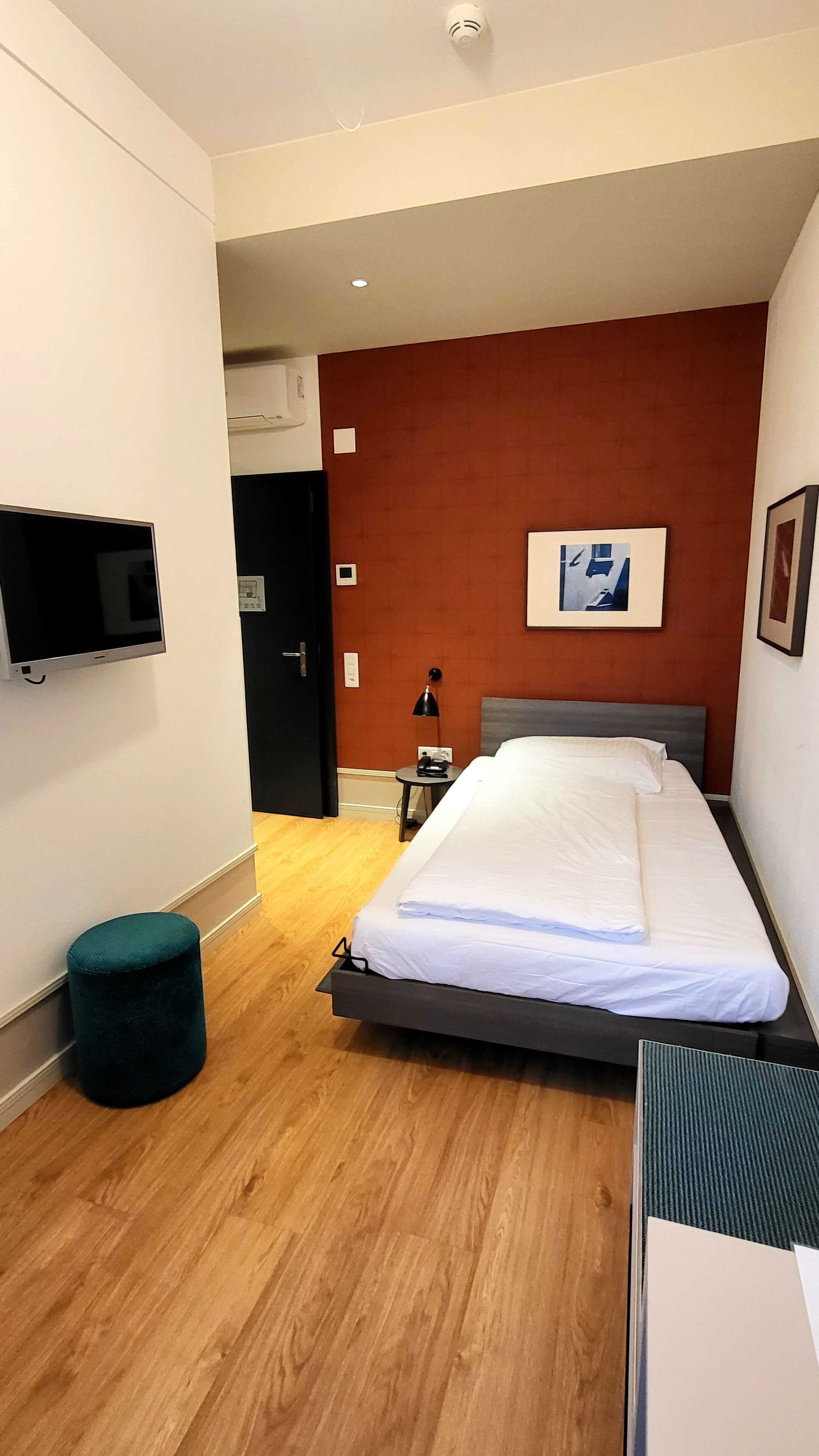A modern hotel room with a bed, a television mounted on the wall, a small table with a lamp, and a door leading to another room.