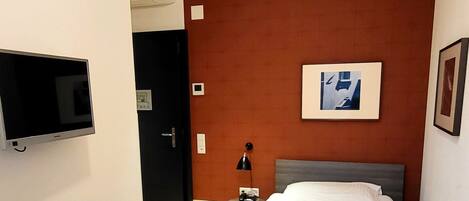 Una habitación de hotel moderna con una cama, un televisor montado en la pared, una mesita de noche con lámpara y una puerta que da a otra habitación.