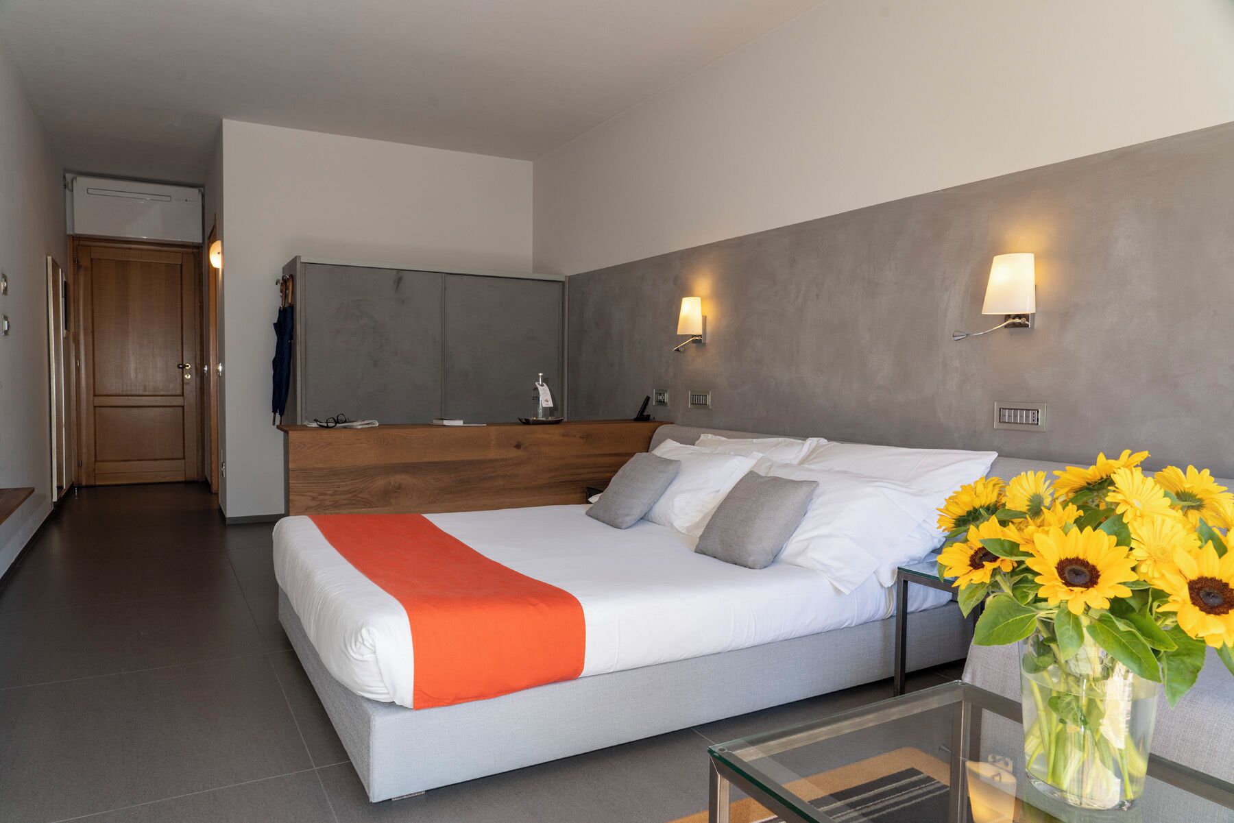 Quarto de hotel moderno com uma cama, uma mesa pequena, um vaso com girassóis e uma cozinha compacta ao fundo.