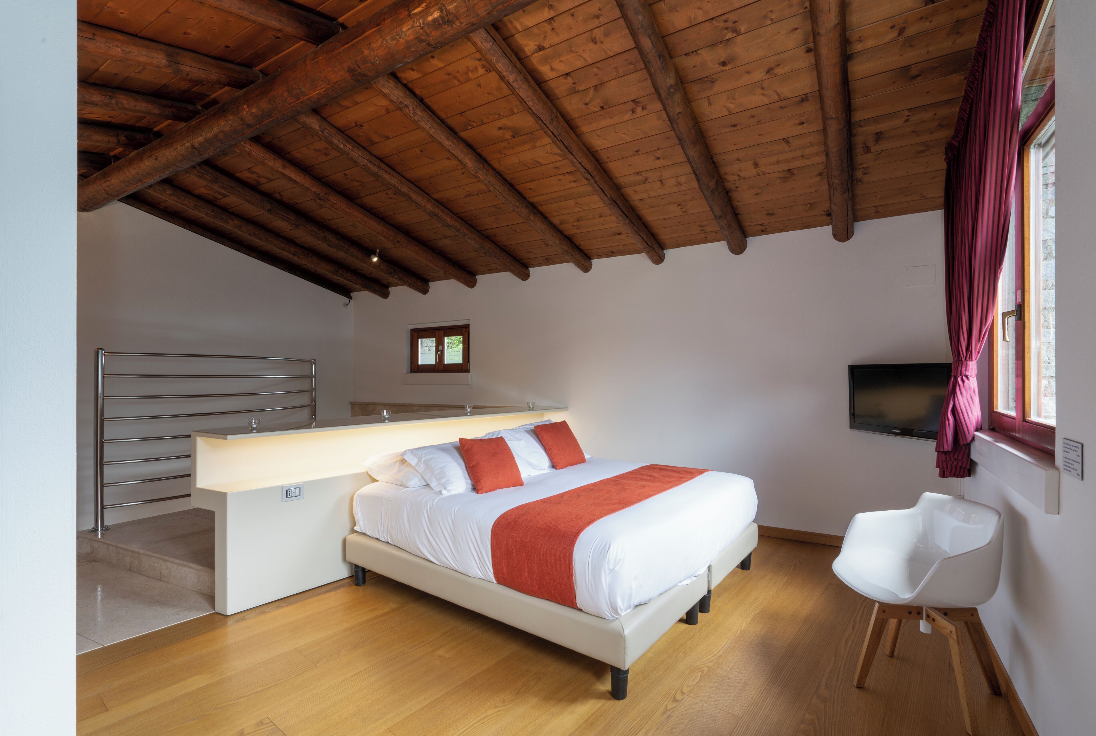 Une chambre avec un plafond en bois, un lit avec une literie blanche et des touches de rouge, une table de chevet, un fauteuil moderne et un téléviseur fixé au mur.
