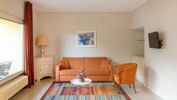 Una sala de estar con sofá, una silla, una mesita de centro, una lámpara, un cuadro en la pared y una ventana con cortinas.