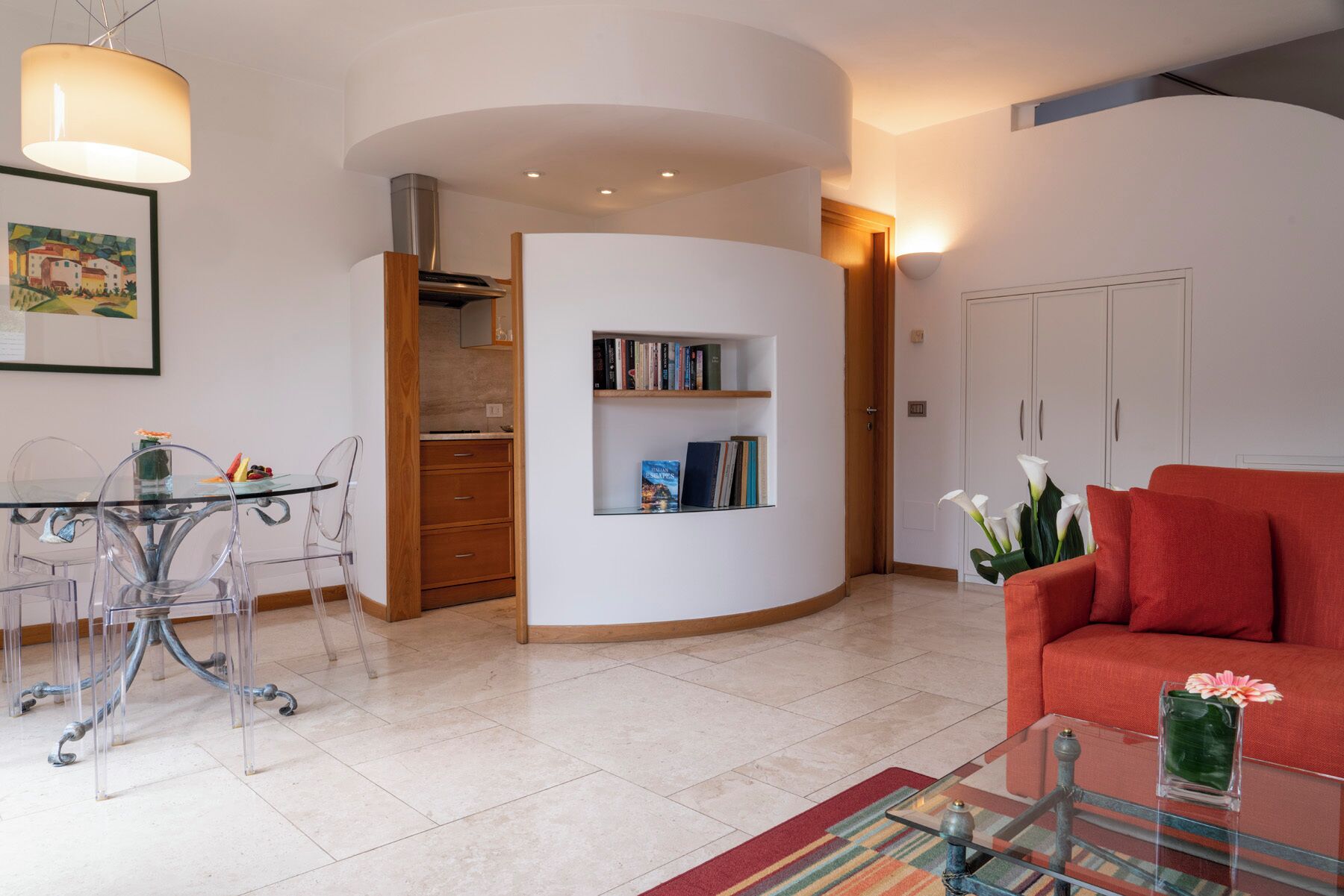 A modern living room with a glass coffee table, a red sofa, a dining area with a glass table and chairs, and a kitchen area with a white cabinet and a bookshelf.