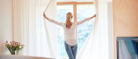 A person standing in a room with a large window, curtains, a flat-screen TV, and a vase with flowers.