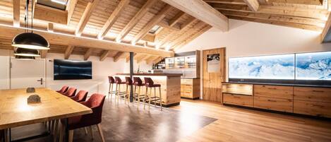 Una cocina moderna con vigas de madera, una zona de comedor con sillas rojas y un televisor grande que muestra una vista de una montaña nevada.