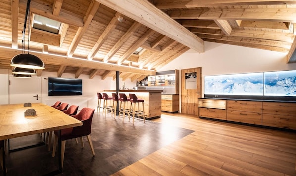 A modern kitchen with wooden beams, a dining area with red chairs, and a large TV displaying a snowy mountain view. - Alpenhotel Schlüssel (Andermatt)