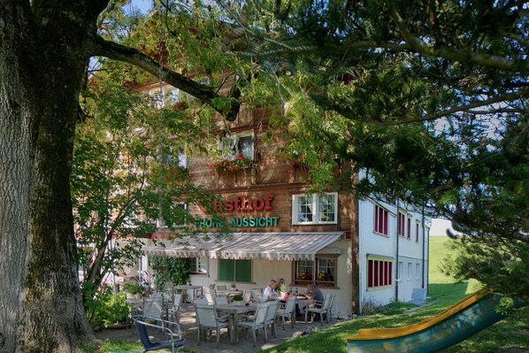 Un restaurante con terraza, un porche cubierto y vistas a una colina.