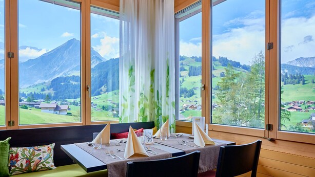 A dining area with a table set for four, large windows with a mountain view, and a bench with a colorful cushion. - Ferien- und Familienhotel Alpina (Adelboden)