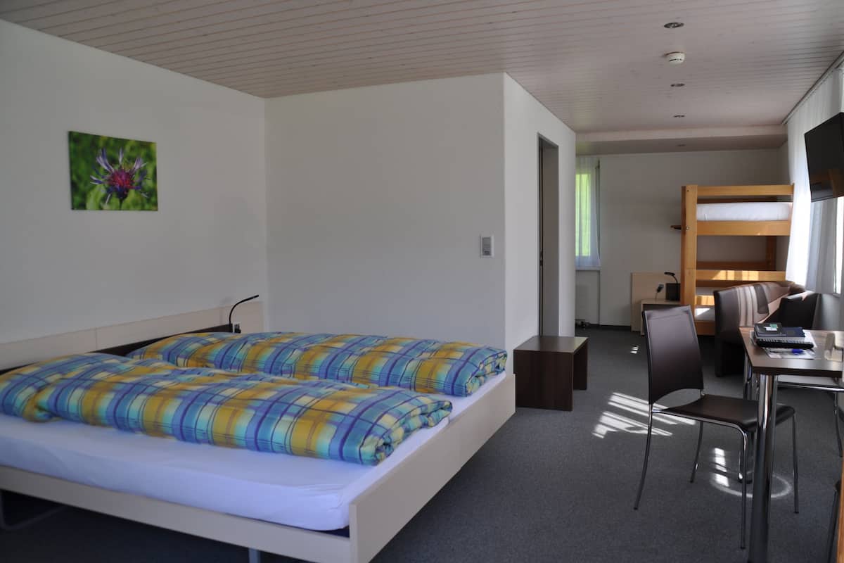A room with a bed, a desk, and a chair. There is a window with curtains and a door leading to another room.