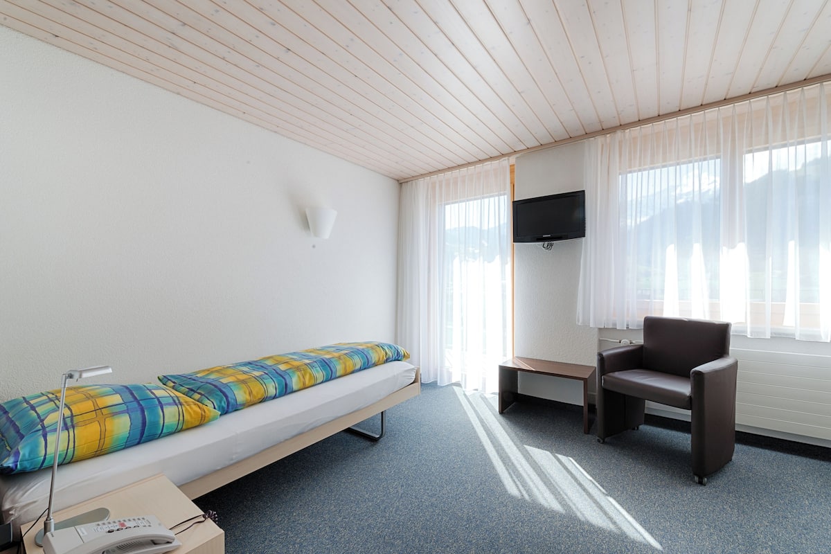 A small, well-lit room with a single bed, a chair, a TV mounted on the wall, and a window with sheer curtains.