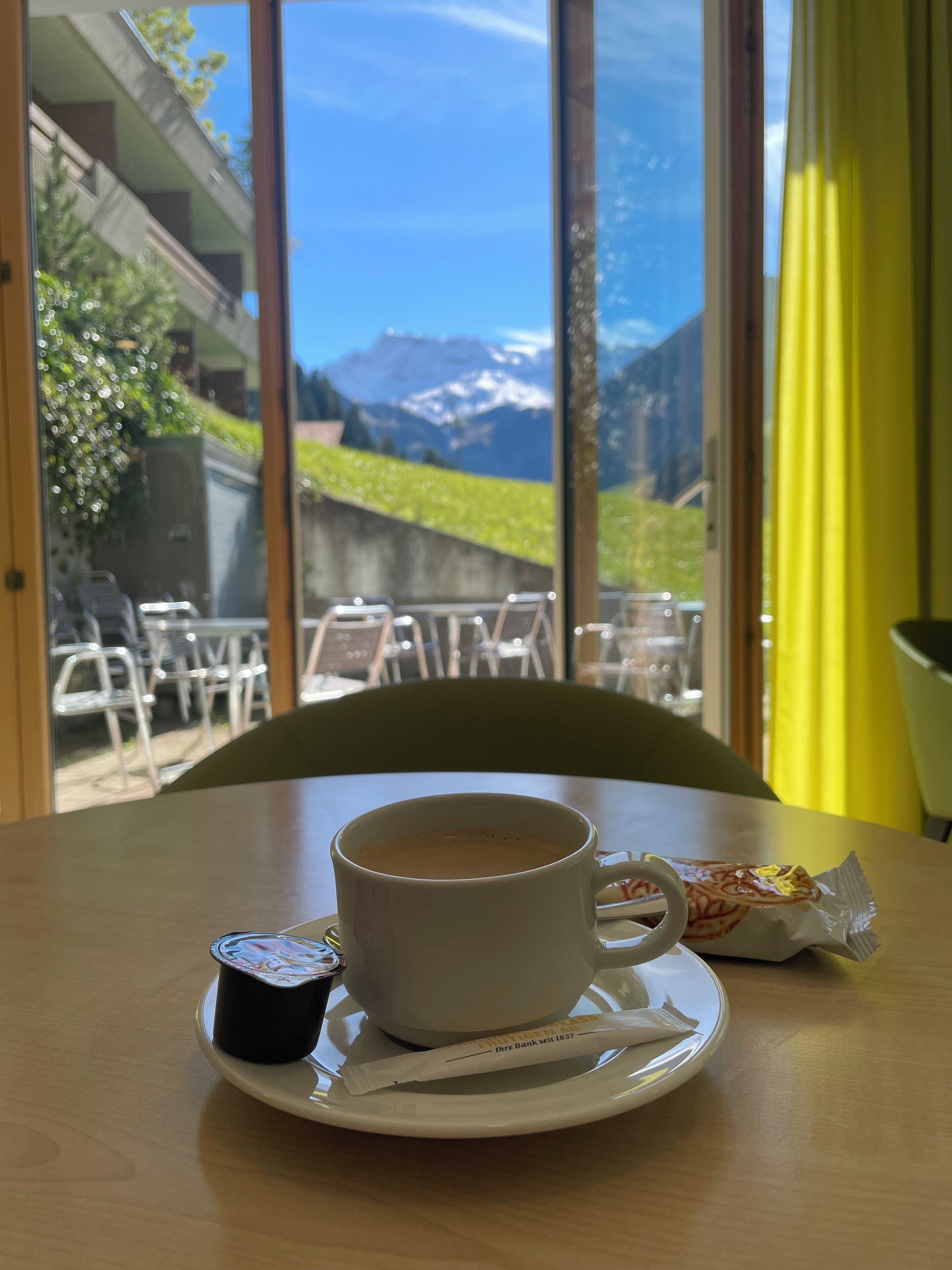 Un tavolo con una tazza di caffè, un pasticcino e una bustina di tè. Il tavolo è vicino a una finestra che si affaccia su montagne e vegetazione.