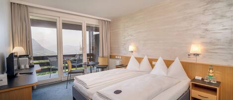 Habitación de hotel con dos camas, un escritorio con ordenador, una silla, una mesita y vistas a las montañas a través de amplias ventanas.