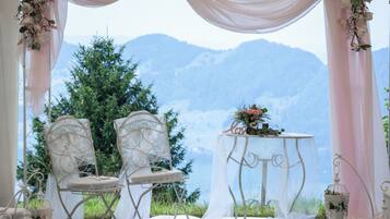 An outdoor wedding setup with two chairs and a table, surrounded by white drapery and floral decorations, set against a mountainous backdrop.
