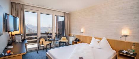 A hotel room with a bed, a desk, a TV, a chair, a table, and a view of mountains through the window.
