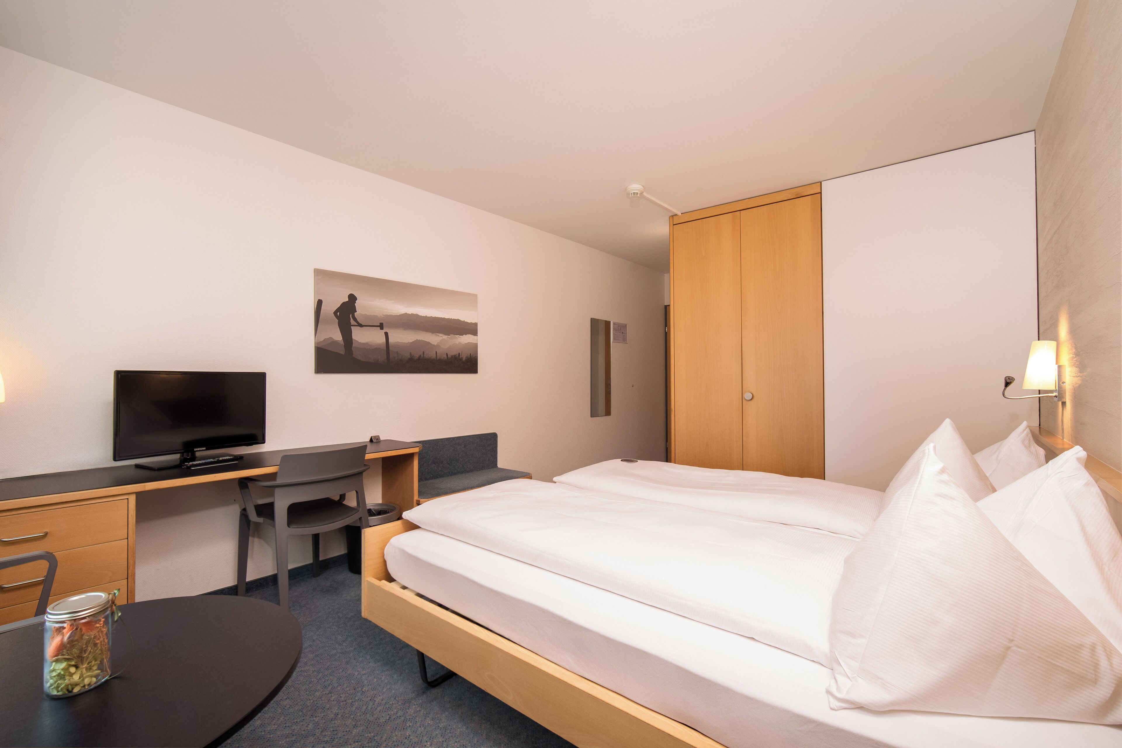 A hotel room with a bed, a desk with a computer, a television, and a painting on the wall.