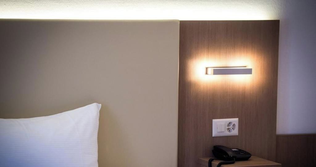 A hotel room with a bedside table, a lamp, and a wall-mounted light fixture.