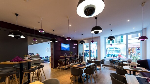 Un café moderno con mesas y sillas de madera, iluminación colgante y una pantalla de televisión en la pared.