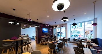 Un café moderno con mesas y sillas de madera, iluminación colgante y una pantalla de televisión en la pared.