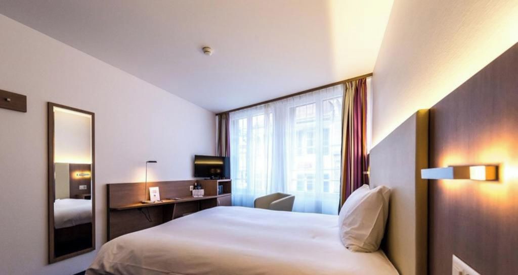 Habitación de hotel con cama, escritorio, silla, televisor y ventana con cortinas.