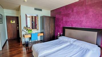A hotel room with a wooden floor, a bed with white bedding, a desk with a chair, a mirror, and a wardrobe.